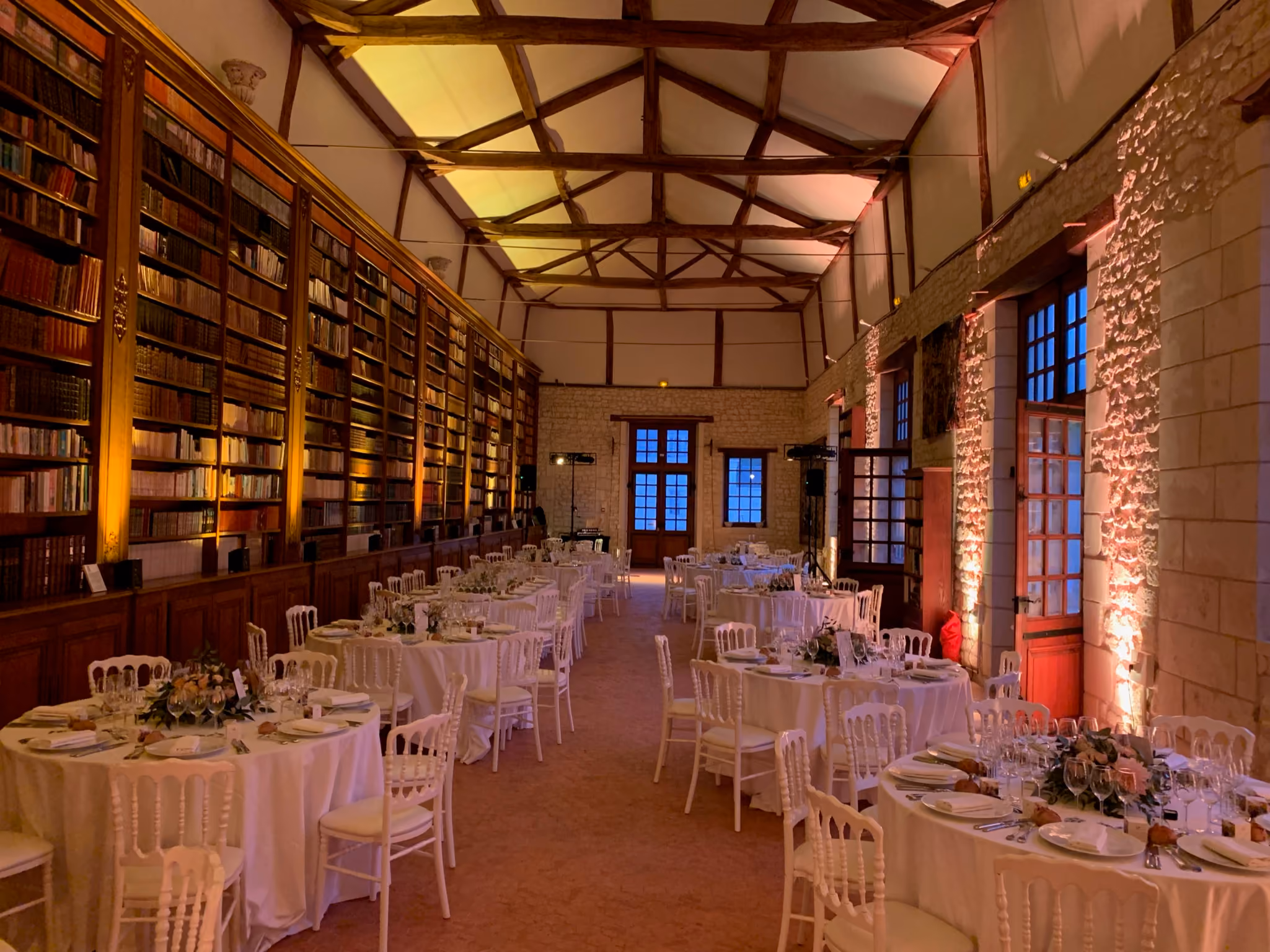 Grande salle bibliothèque dressée en tables rondes sous une charpente apparente éclairée en ambre