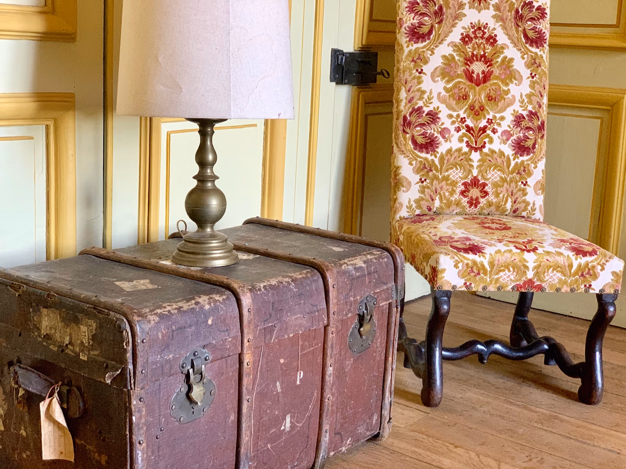 Malle de voyage ancienne et chaise en velours damassé devant une porte à boiseries dorées